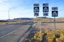 Road signs off a rural street in an arid area. There are a few buildings in the distance and mountains beyond that.