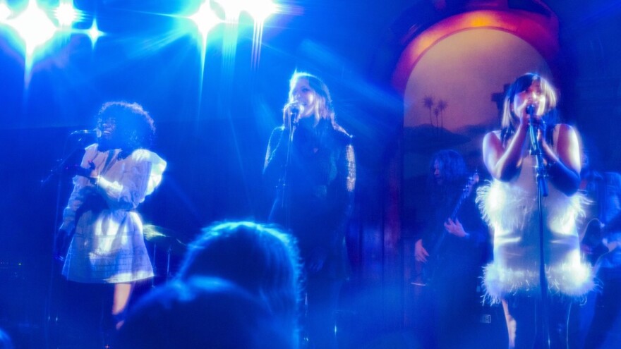  Three women in fashionable dresses sing into standing microphones on stage with bright lights overhead.