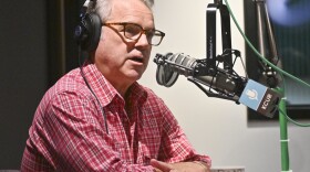 A man sitting in a radio studio sits behind a microphone and is talking.