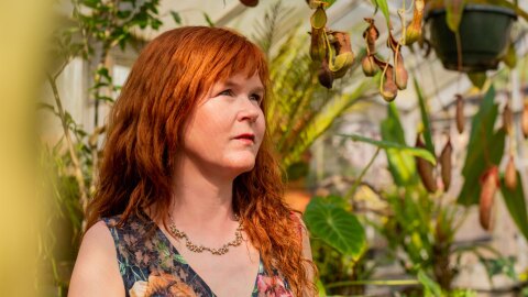 Pianist Sarah Cahill stands in a lush greenhouse filled with various vibrant plants, including a hanging pot of tropical pitcher plants. Sunlight filters through the glass structure, illuminating the scene.