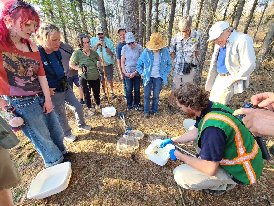 people of various ages stand in a semi-circle around a man in green vest with a tub of water and salamanders