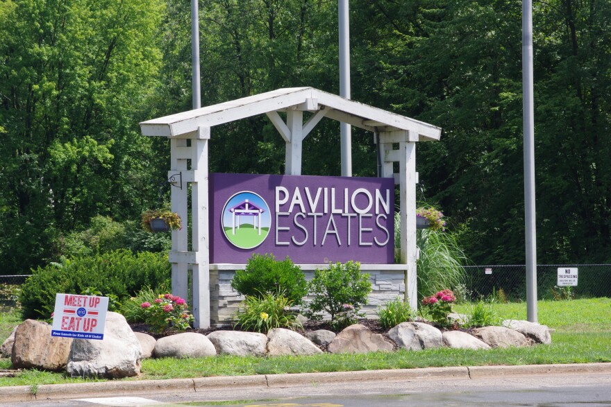 A large white and purple sign reads "Pavilion Estate." The sign is surrounded by flowers, bushes and rocks. A road passes just in front of it, with a small sign on a fence in the background reading "no trespassing."