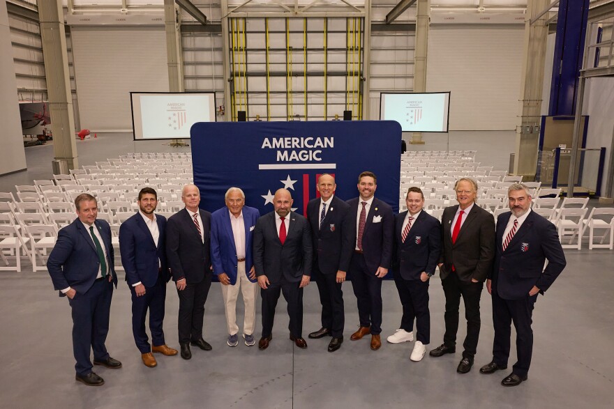 Group of American Magic and SailGP representatives standing with city and state officials inside a large industrial facility at the Port of Pensacola, with American Magic branding and rows of white chairs behind them.