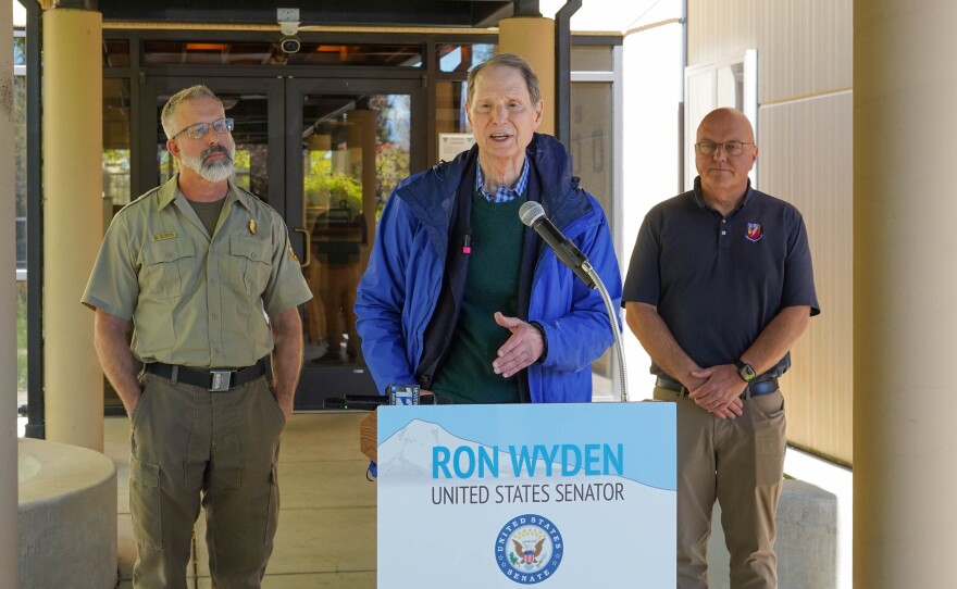 Senator Ron Wyden speaking in front of a podium with his name. Two men stand on either side.