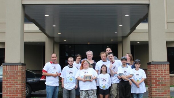 A dozen caregivers and clients stand in front of the Access CNY's brick and stucco building smiling while they hold an award recognizing its Self-Service Advocacy Club. 