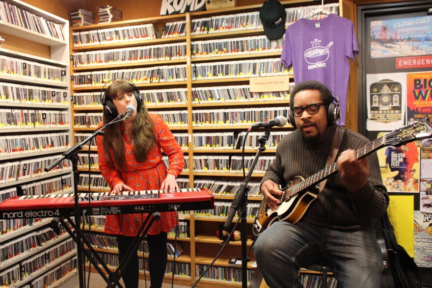 Woman in a red dress standing playing a red keyboard and singing into a microphone, man in a brown sweater seated playing bass