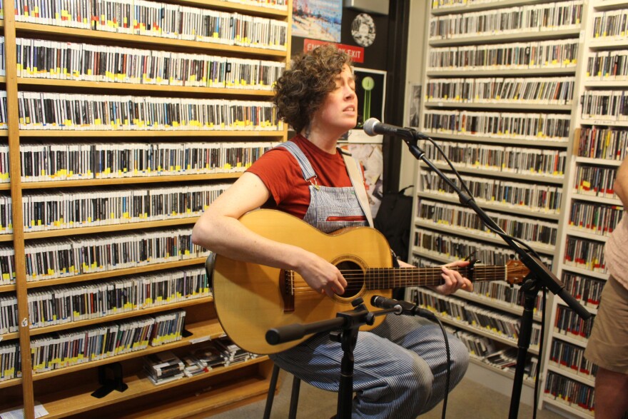 A woman sits on a stool in a radio studio that has shelves of CDs lining the walls and sings into a microphone while playing guitar.