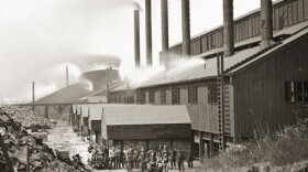Exterior of the Bay View Rolling Mill, Puddling Department, with workers posing outside the building. The men are standing near railroad tracks, and a few of the men are sitting on a small railroad car.