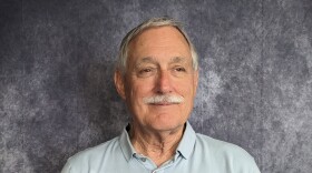 A white-haired man in a light blue shirt smiles slightly.