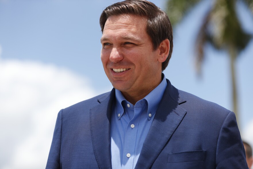 DeSantis looks off to the side while smiling. He's wearing a button-down shirt and blazer. Behind him is one palm tree.