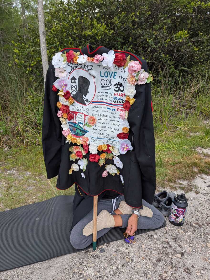 A man sits cross-legged while holding a uniform with a sign pasted on that says "STOP ICE"