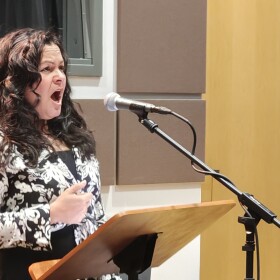 Singer Cecilia Duarte performs at a microphone, standing at a music stand in a recital hall.