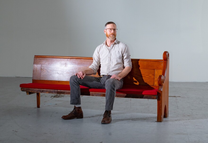 A bearded man wearing glasses, jeans and a button-down shirt sits on a broken church pew in an otherwise-empty room.