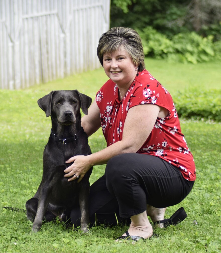 A white woman in a red shirt kneels next to a sitting dog on a lawn