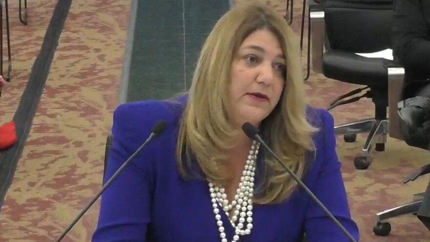 Woman sits at table in purple suit with pearls in front of microphones