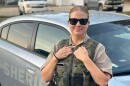 A woman is wearing a law enforcement uniform, sunglasses and is standing in front of a sheriff's car.
