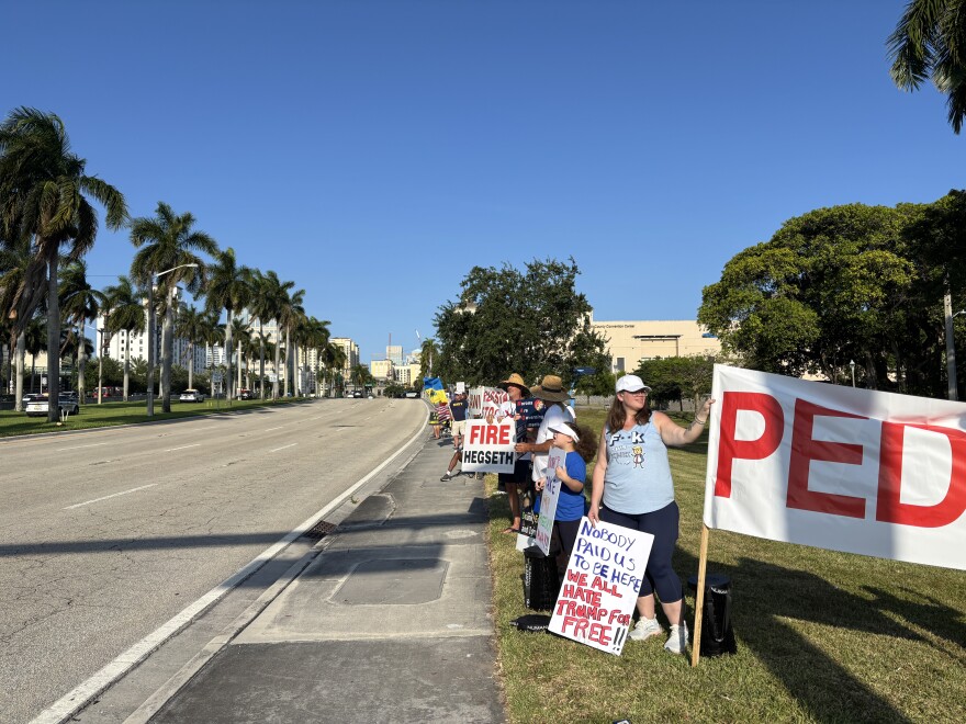Protestors hold large-lettered signs for cars to see during rush hour traffic on Okeechobee Boulevard in West Palm Beach, near the Kravis Center where President Trump is giving a speech on Friday, May 1, 2026.