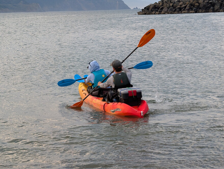 two people sit in a two-person orange kayak paddling out to sea. They have a box attached to the back of the kayak