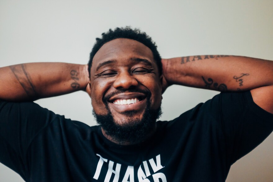 A Black man in a black shirt smiling. He has his hands behind his head 