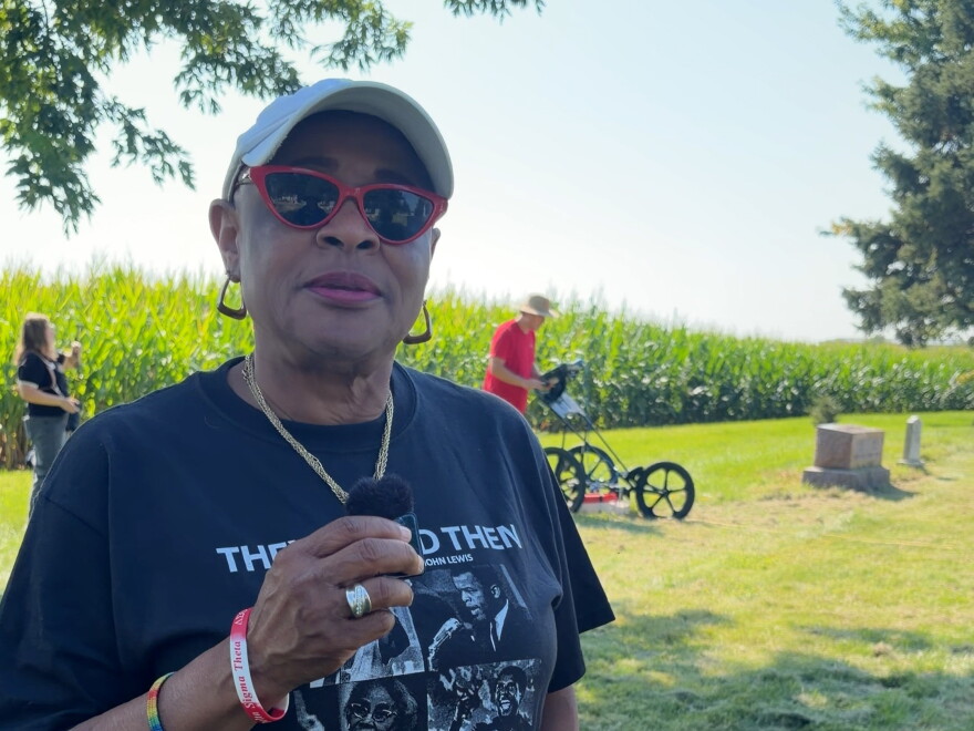 a woman wearing sunglasses and a baseball cap stands in front of a row of corn and green grass