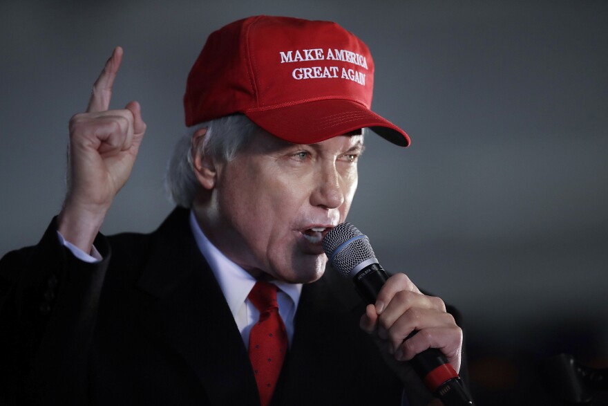 Three-quarter head-and-shoulders view of a man with white hair wearing a dark suit with white shirt, red tie and red "Make America Great Again" hat, as he speaks into a microphone while gesturing with his right index finger raised