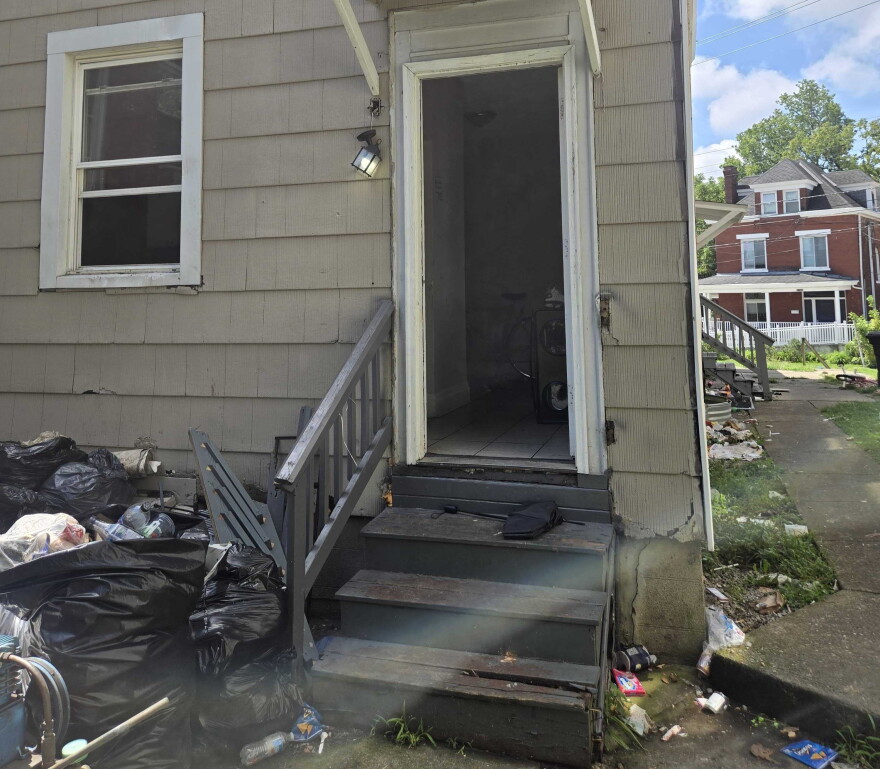 A rear entrance to a single family home. The door and a window are wide open and trash is piled on the ground.
