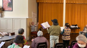 A woman sits at a grand piano and gestures to a choir of people sitting in chairs during a rehearsal