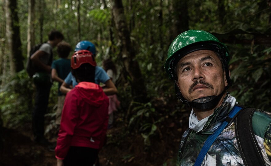 A man wearing a helmet and outdoor gear looks in the distance while facing the camera, as a line of people wearing raincoats and helmets walk through the jungle behind him.