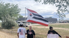 A boy in an American flag t-shirt holds up an Arapaho flag. He stands next to an older woman in a black t-shirt and another woman in a colorful skirt holding up a protest sign.