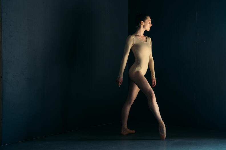 A woman wearing a tan leotard in front of a dark black background. She is standing with her arms by her side and her right leg pointed.