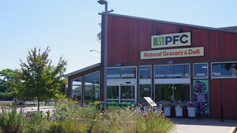 a red building with a slanted roof sits in front of thin flowers, weeds and other plants on a concrete walking path. The sign for the store reads "PFC Natural Grocery & Deli."