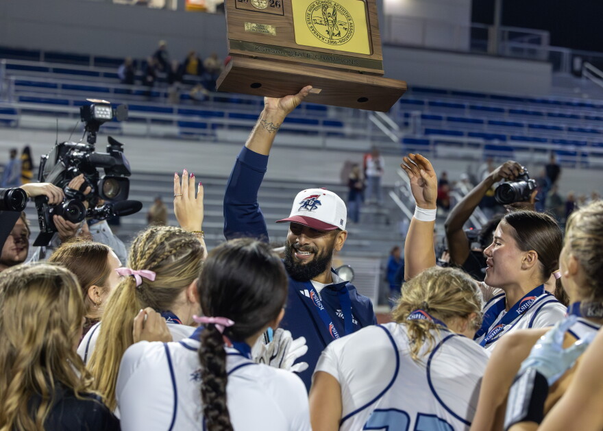 Coach Matt Hightower is lifting the state championship plaque overhead, surrounded by the team with cameras pointing in to the huddle. He has a beard and is wearing a white hat with the school's logo. He's facing toward the camera with a large smile.