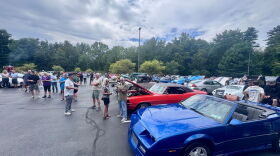 There's rows of muscle cars in a parking lot, which a bunch of people standing around talking. The sky is mostly cloudy, and trees line the parking lot. 