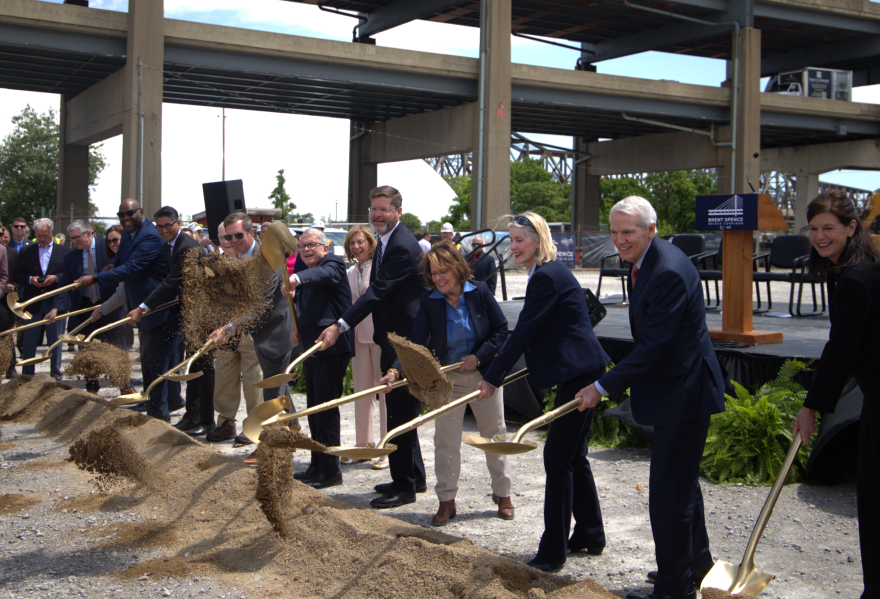 A group of people in dress clothes shovel dirt next to a large steel bridge