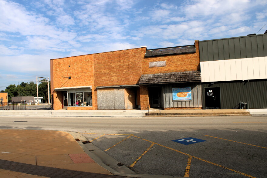 A street view of old brick and modern buildings, one with boarded-up windows. A storefront has a faded sign and a blue handicapped parking space is marked in the foreground. The sky is partly cloudy.
