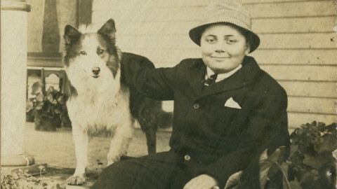 A monochromatic photograph of a young Herman B Wells wearing a dark suit jacket and a brimmed hat, seated outdoors while resting his arm on his standing, long-haired collie dog.