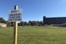 in the foreground, a white sign on a wooden post. in the background, an open field and brown brick building