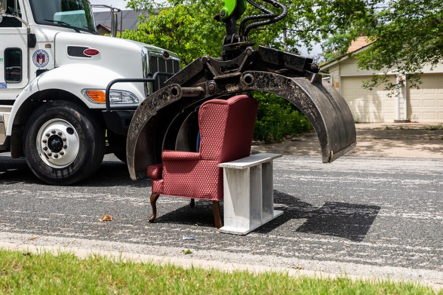 A big metal claw reaches down to pick up an upholstered chair and shelf from the street 