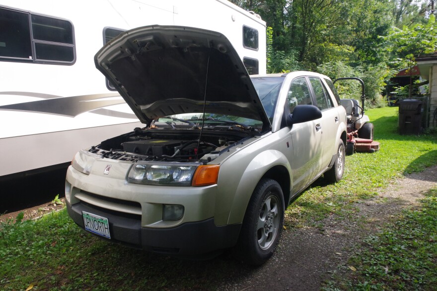 a light tan SUV with the hood open is in the foreground. Behind it is a riding mower.