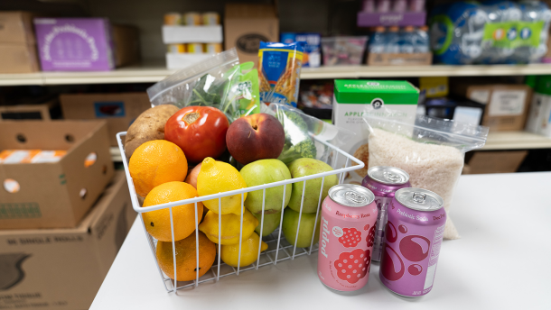 Photo of a white grocery basket with oranges, lemons, green apples, tomatoes, peaches and potatoes. Beside it are canned drinks and a bag of rice.