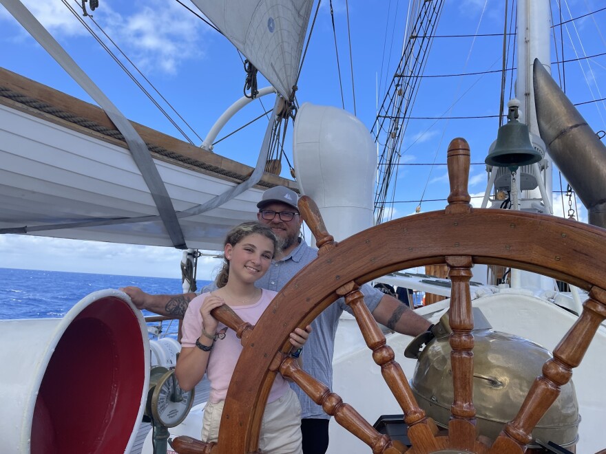 A man and a girl stand behind the wheel of a tall ship and smile at the camera. 