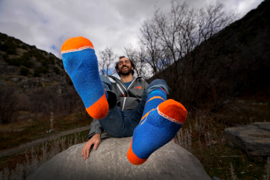 Jack Danos sits on a rock while holding up his feet to show a pair of blue and orange ColdSlayer socks.