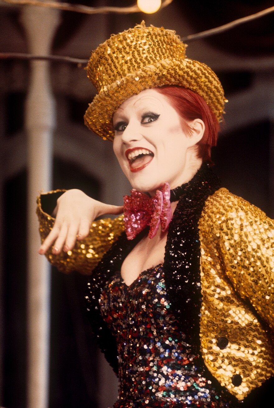 Person wearing a glittery gold jacket and hat, with bright red hair and heavy makeup. They are smiling and striking a playful pose, with a sequined top and a pink bowtie under stage lights.