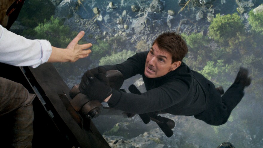 Actor Tom Cruise hangs from the side of a helicopter with the ground several hundred feet below him.