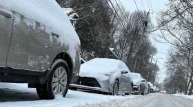 a line of snow-covered cars along a neighborhood street in Syracuse with one car driving with headlights on.