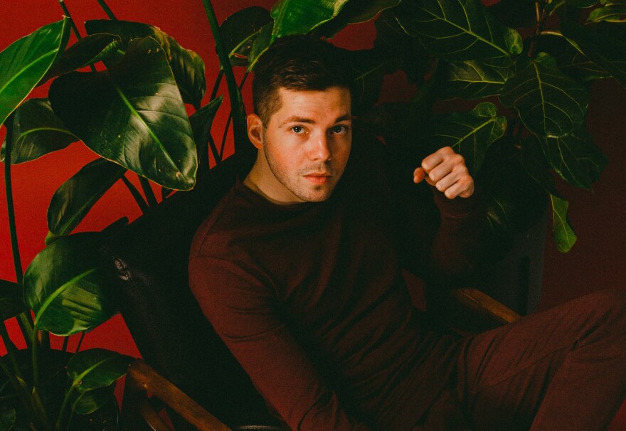 Danny Arcane sits on a leather chair surrounded by plants in front of a red background.