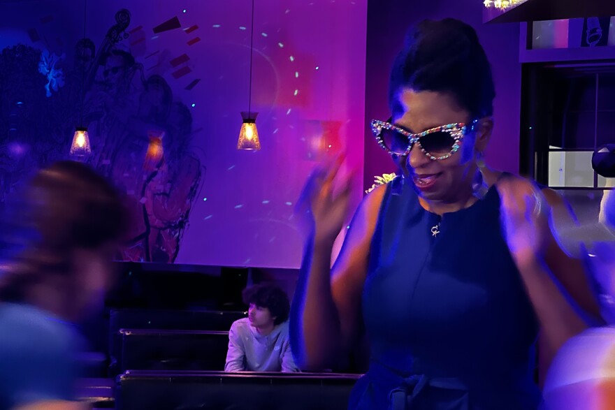 McCray wears oversized, bedazzled sunglasses on a dance floor illuminated in purple and blue lights. 