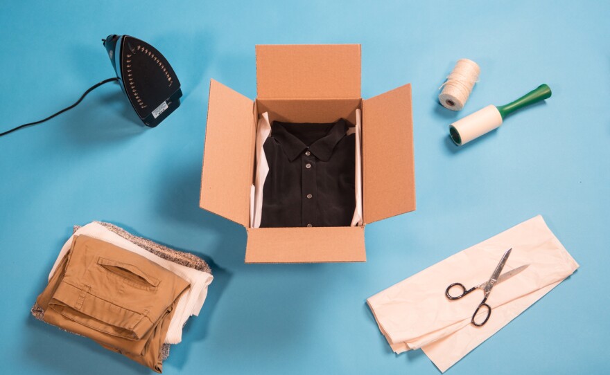 A black collared shirt is neatly folded inside a cardboard box on a blue background in the middle of the frame. The box is surrounded by an iron, a stack of clothes, a pair of scissors, tissue paper, twine and a lint roller.