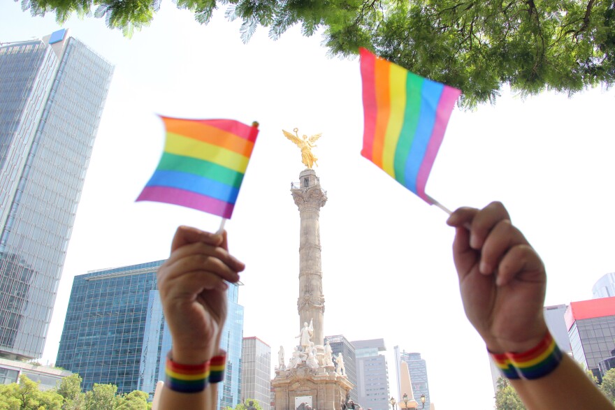 Two hands hold miniature rainbow flags in front of the Angel of Independence statue in Mexico City