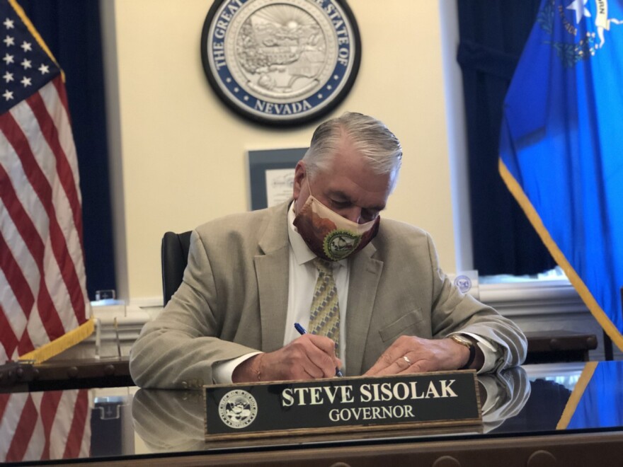 Nevada Governor Steve Sisolak is sitting at a desk and signing a bill. He is looking down at the bill and wearing a face mask. There is a name plaque in front of him on the desk, and the seal of Nevada is visible on the wall behind him.